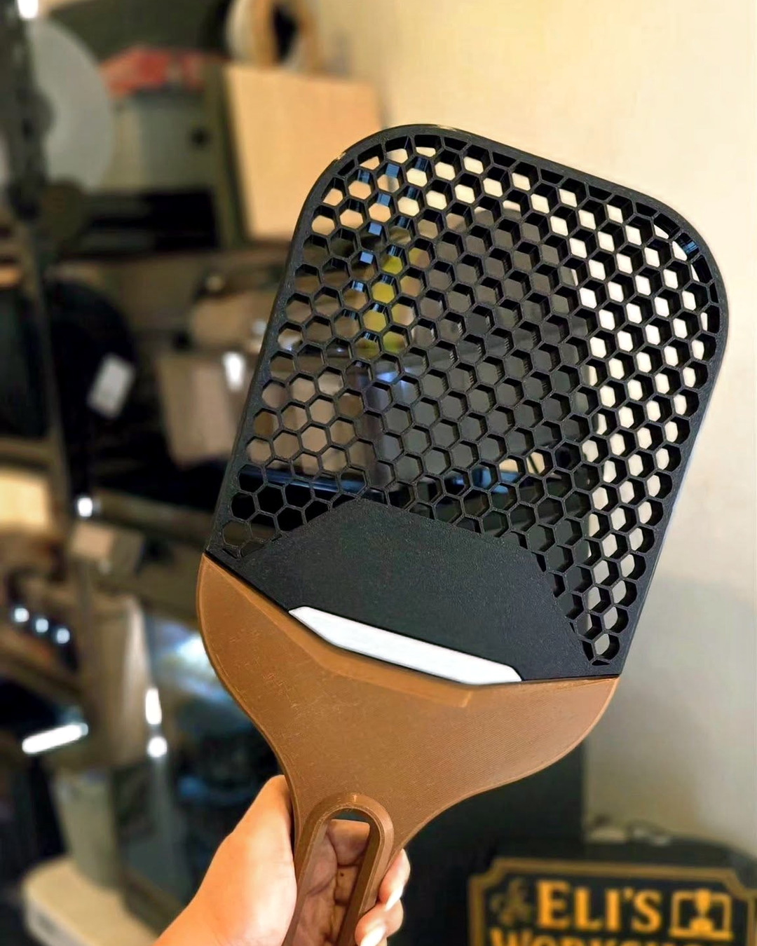 A honeycomb-mesh paddle in brown, black and white, held up by hand next to the Eli's Workshop sign.